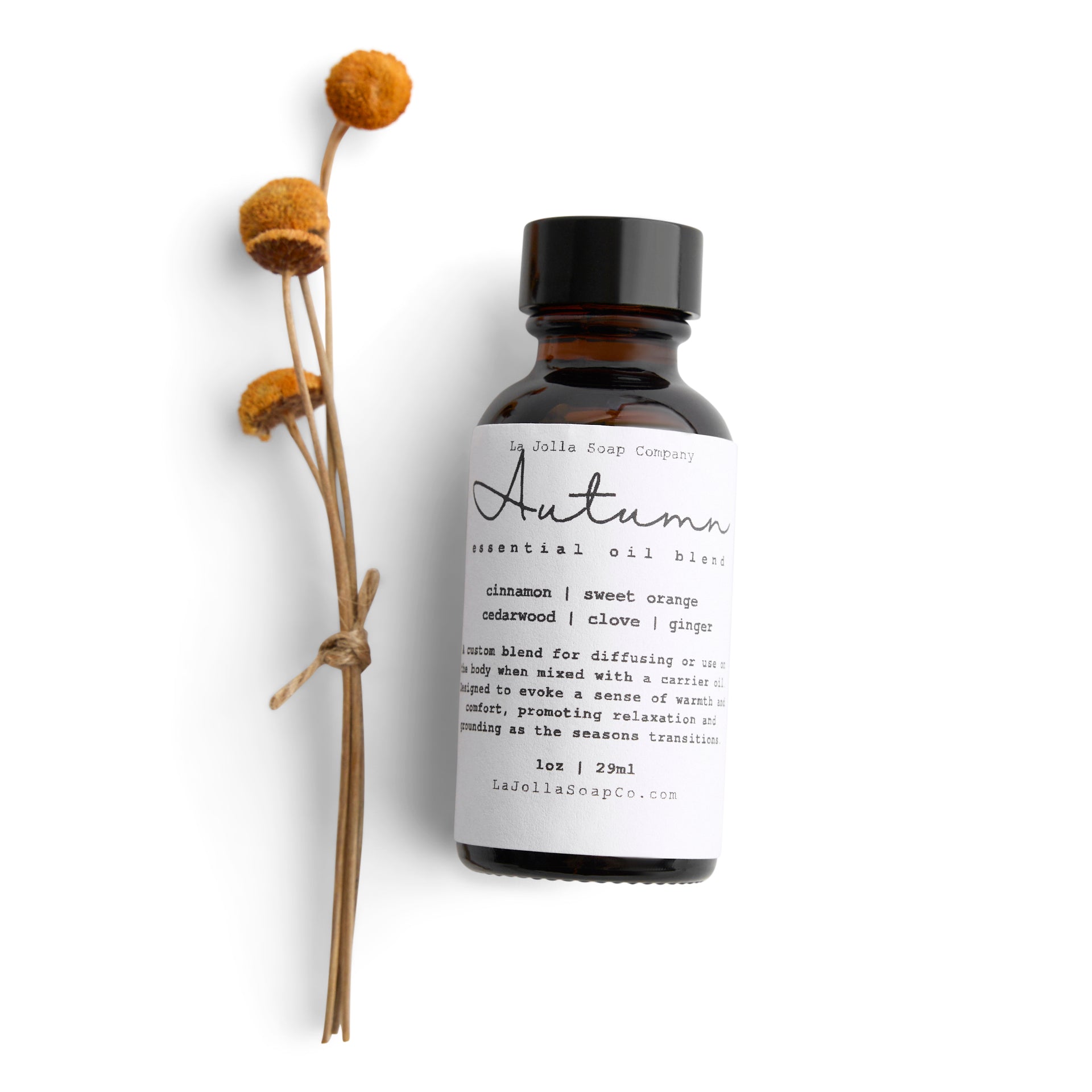 A bottle of Autumn essential oil blend with small orange Billy button flowers laid in front. The blend evokes a warm and cozy seasonal vibe.