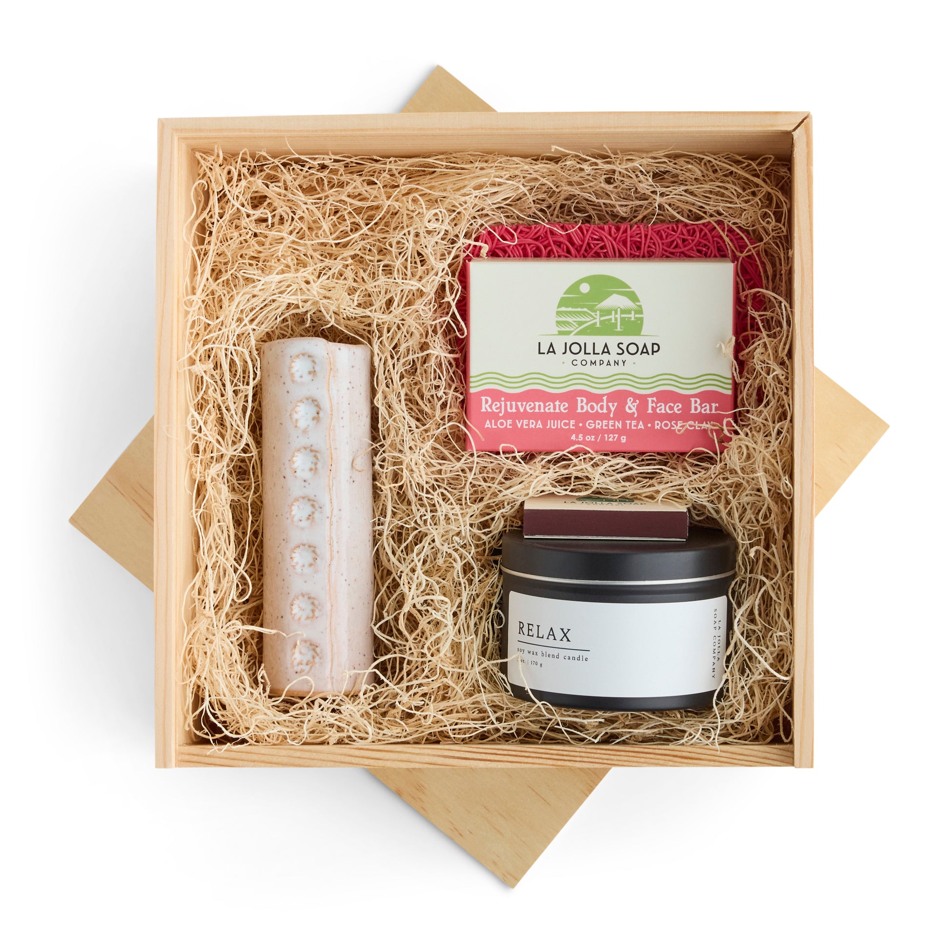 Relax and Rejuvenate gift box featuring a Rejuvenate body and face bar on a pink soap rest, a white ceramic vase with white button flowers, a Relax 6 oz. candle ian a black tin, and a book of La Jolla Soap company matches. Presented in a natural pine wood keepsake box.