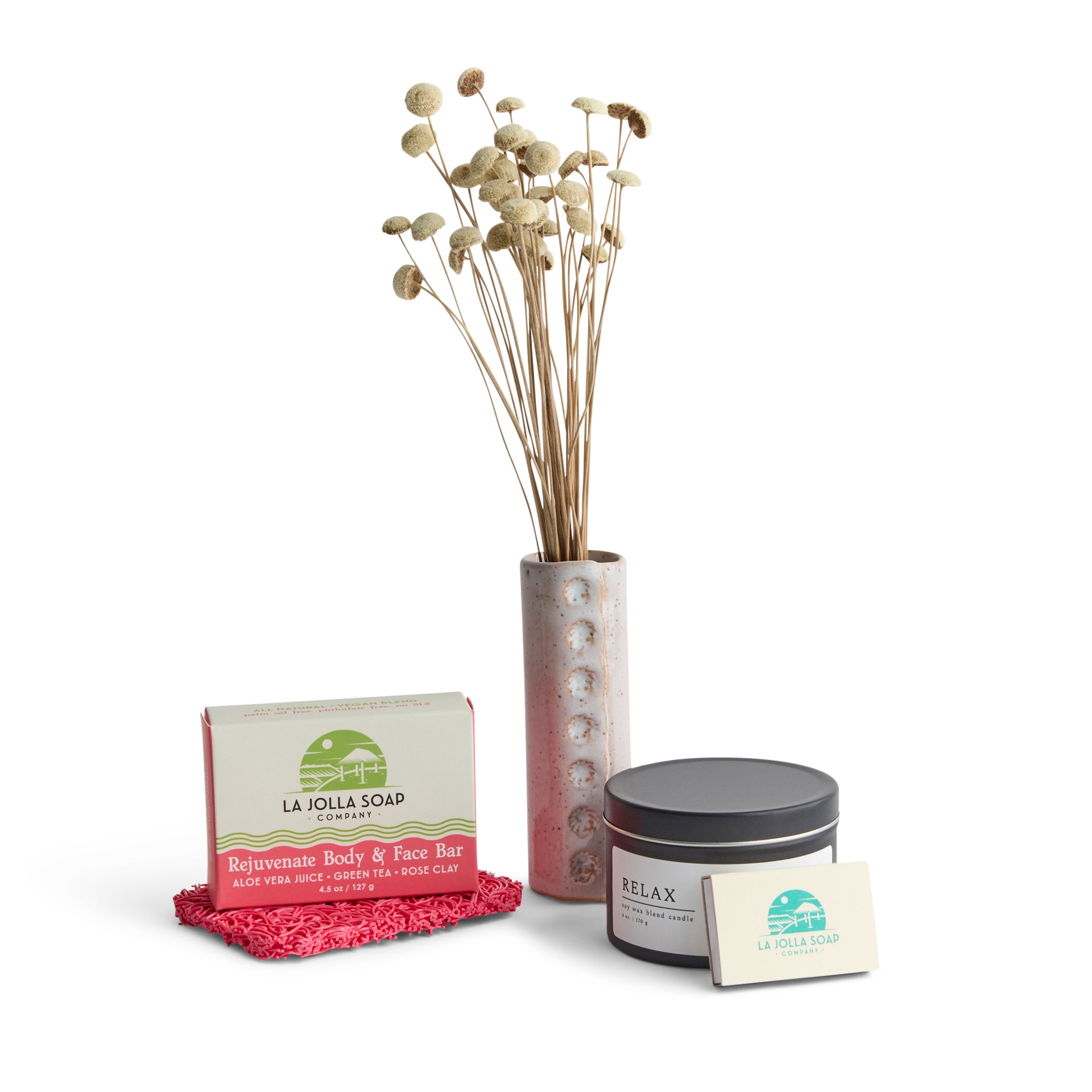 Rejuvenate and relax set featuring a Rejuvenate body and face soap bar resting on a pink soap rest, accompanied by a white ceramic vase with delicate white button flowers, a Relax 6 oz. candle in a black tin, and a book of La Jolla Soap Company matches.