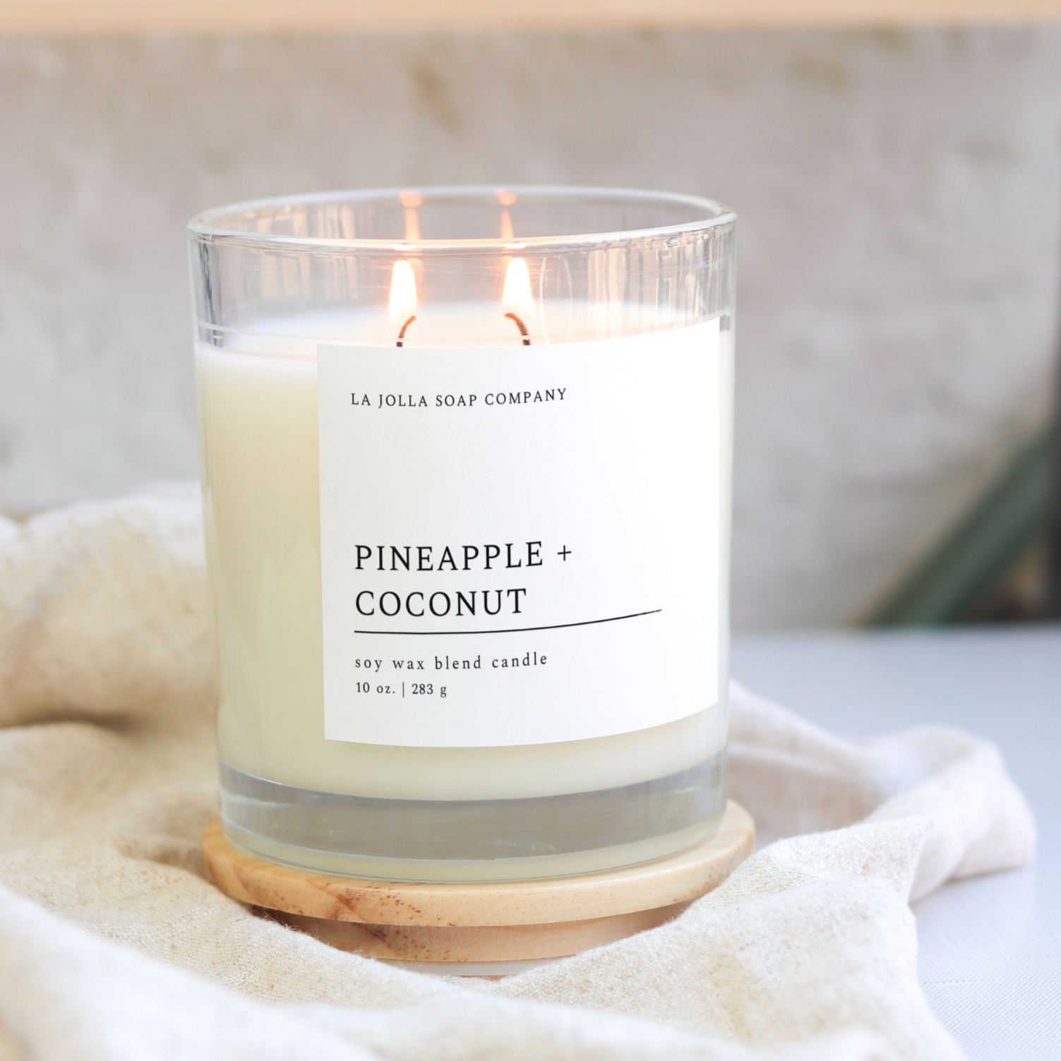 Pineapple + Coconut 10 oz candle in a glass jar with  wood lid. Pineapple and coconut, blended together in perfect harmony. The aroma conjures images of swaying palm trees and sun-drenched shores, offering a serene Tropical Paradise in your own home.