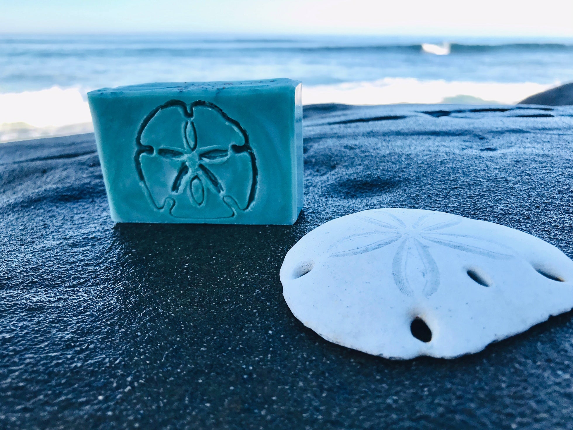 La Jolla Soap Company's Sea Clay Soap. The soap is turquoise blue with a sand dollar carved on the face of the soap. There is a sand dollar laying next to it. The ocean waves are crashing behind them.