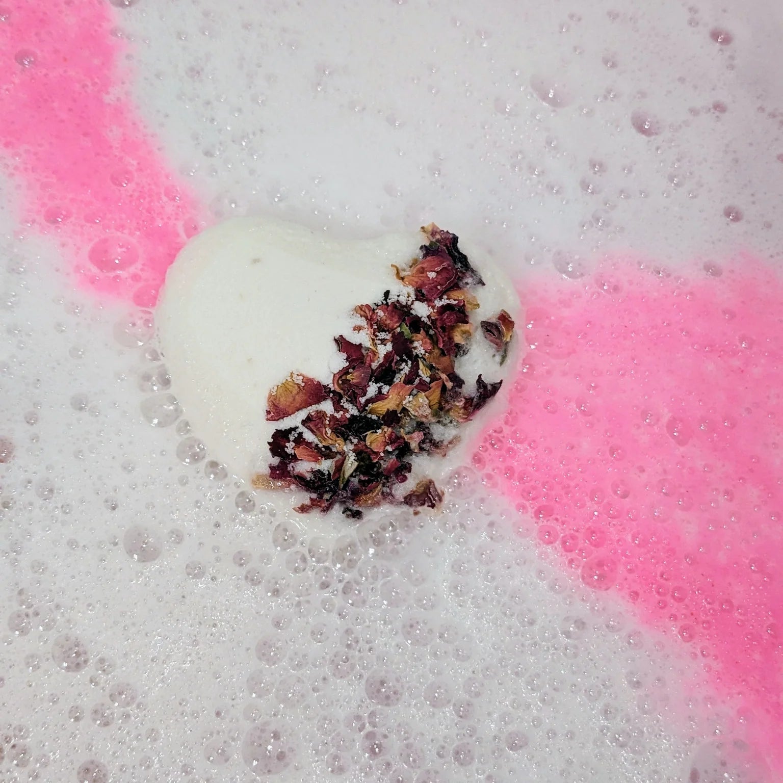 White heart-shaped bath bomb topped with delicate red rose petals, fizzing in water, streaming pink frothy foam, transforming the bathwater into a dreamy shade of pink.