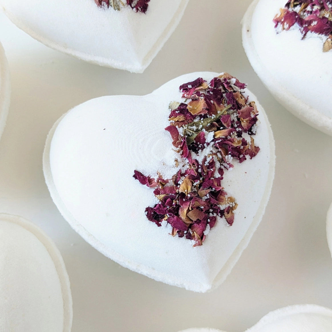 white heart-shaped bath bomb topped with delicate red rose petals, with a natural white background. 