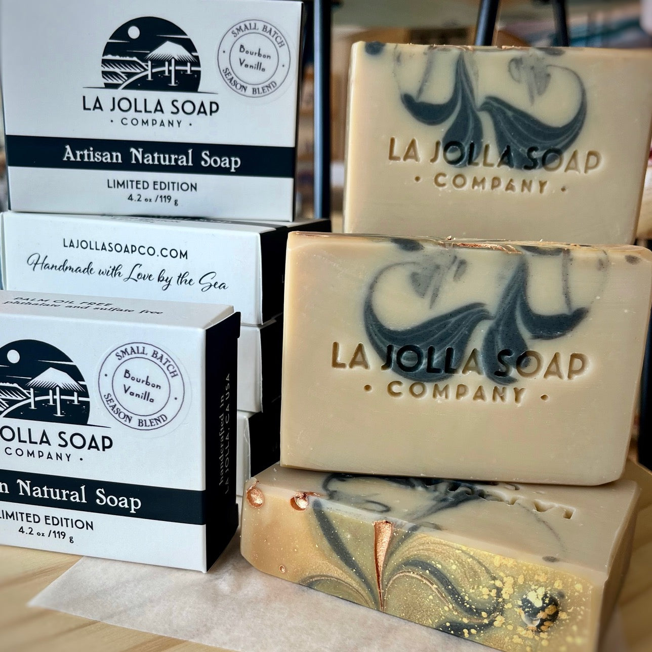 Bourbon vanilla soap, creamy latte color with lovely black swirls, placed on top of a wooden table sitting next to the boxes it is packaged in, white in color with a rich black accent.  