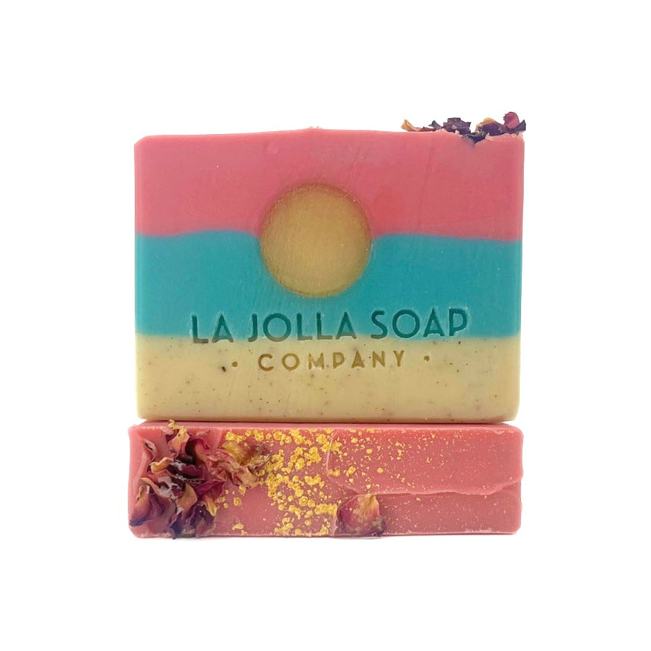 A beautifully handcrafted soap bar with three distinct layers, a creamy sand-colored base, a vibrant tropical ocean blue middle, and a rich dark pink top. a golden sun design sits at the center, while the top is adorned with shimmering gold accents and delicate dried rose petals. Part of the For Love and Roses Collection, his artisan soap evokes the warmth of a tropical sunrise